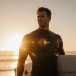 Chris Hemsworth carrying surf wax on sunrise shore